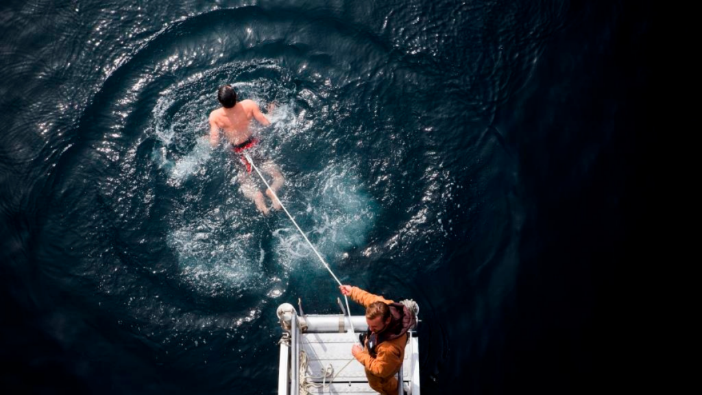 Man tethered to rope swimming in Arctic waters with a man holding the rope from a deck